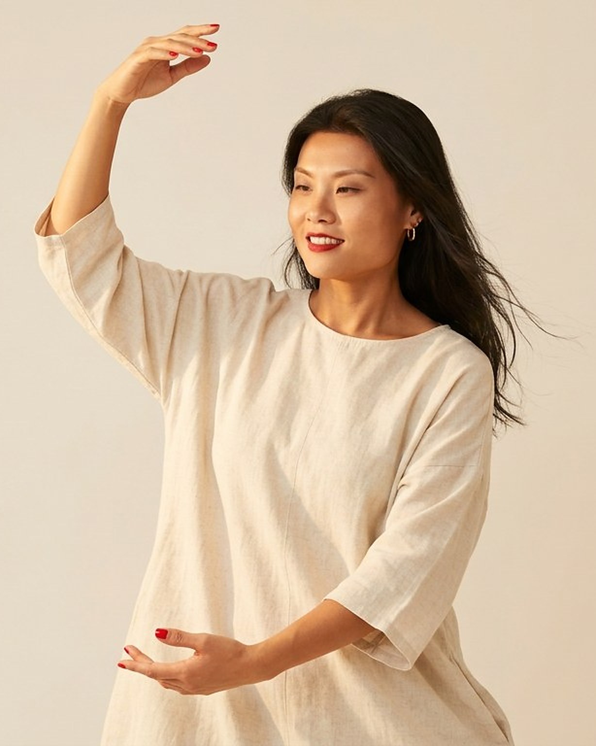Yan Huang holding a slow qigong movement in a loose cream linen tunic against a warm pale backdrop